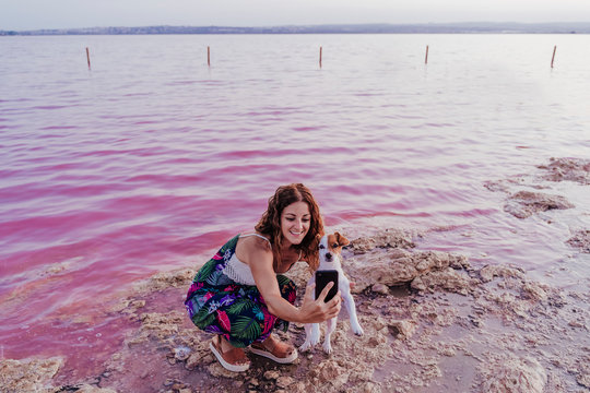 young beautiful woman standing by a pink lake or lagoon in a natural park at sunset. Torrevieja, Spain. Cute jack russell small dog besides.Tourism, love and travel concept