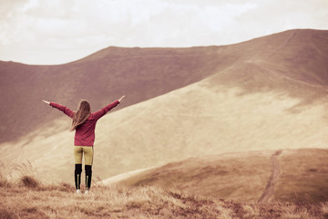 Traveling girl with a raised hands standing in mountains with her hair fluttering in the wind. Toned picture.