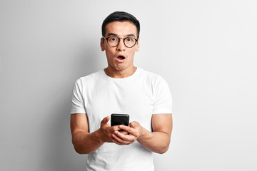 Handsome Asian guy in white studio looks surprised at screen of mobile phone. Shocked Kazakh man in glasses dressed casual holds smartphone in hands