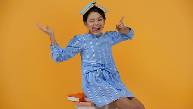 excited schoolgirl with book on head grimacing isolated on yellow