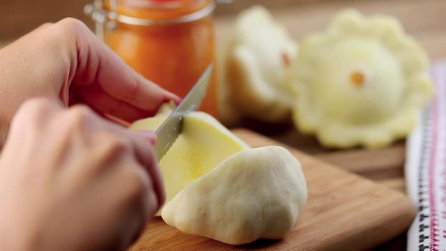 Female hands cutting a small white squash by knife on a wooden cutting board with a squash and glass jar on the background
