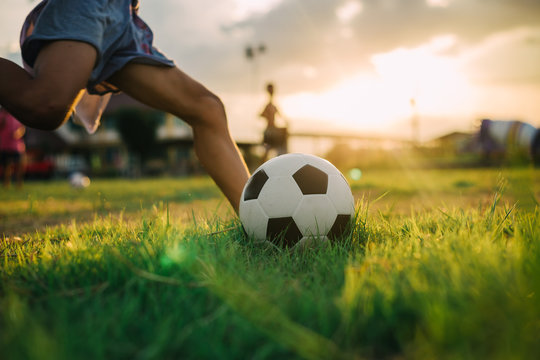 A boy kicking a ball with bare foot while playing street soccer football on the green grass field for exercise in community rural area under the twilight sunset.