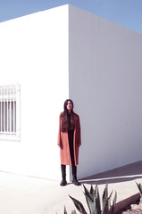 young long-haired woman wears a trench coat and stands in front of a corner of a wall where one...