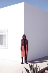 Young long-haired woman wears a trench coat. She is looking at the floor and standing in front of a corner of a wall where one side is lit and another is in the shade