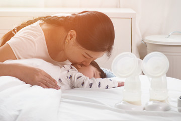Obraz na płótnie Canvas Asian mother touching and kissing her cute newborn sleeping baby after using twin breast pump to feed her child in the bedroom at home. Mother cradling baby with love and baby feeding concept