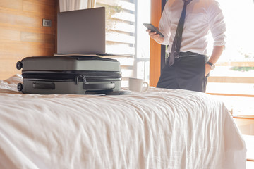 Businessman looking at cell phone with laptop on top of suitcase on the bed.