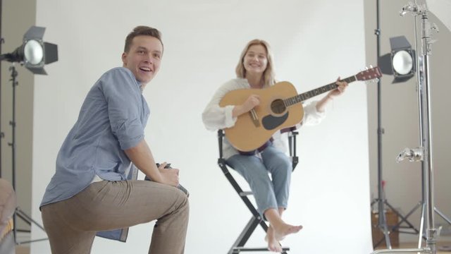 Backstage of the photo shoot. Professional fun photographer taking photos of young girl playing guitar while sitting on the chair on white background in the studio. Fashion studio photoshoot.