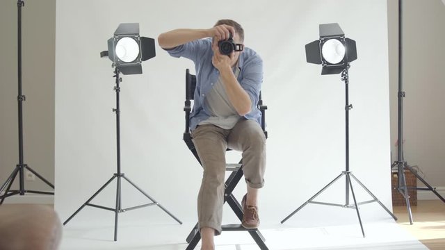 Professional photographer taking photos while sitting on the chair and looking at camera. Photographer on white background in the studio. Fashion magazine studio photoshoot.