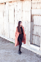 young woman with her hands in the pockets of her trench coat walks pensively in front of an old wooden wall