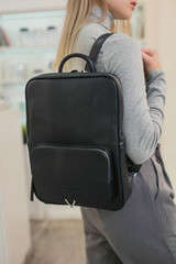 A girl in a gray turtleneck with a leather backpack on her back. Business lady standing in the room with a backpack made of eco-leather.