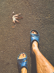 Bare male legs in blue slippers on the asphalt ground. Autumn leaf, the coming of spring