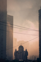 Beautiful scene of morning sunlight through the dense fog between high modern city buildings, and small christian orthodox church silhouette in the middle.