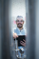 Shot of smiling middle aged caucasian technologist expert with hairnet and protective glasses operating industrial machine in food or pharmaceutical factory.