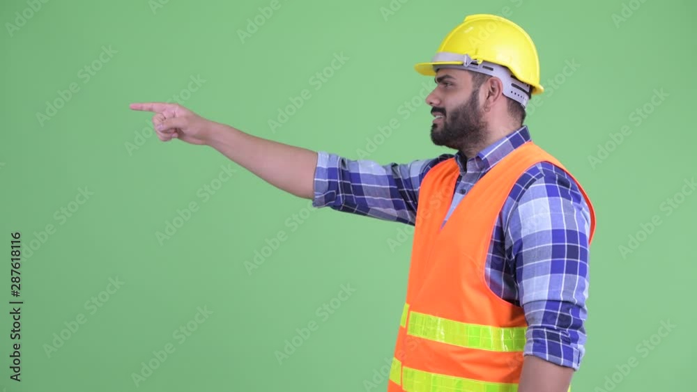 Wall mural Profile view of happy young overweight bearded Indian man construction worker pointing finger