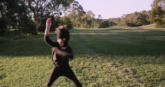 young boy wearing a homemade ninja mask and black costume practices ninja moves, ninjutsu, jumping, kicking and running around a park in the late afternoon golden glow of the setting sun