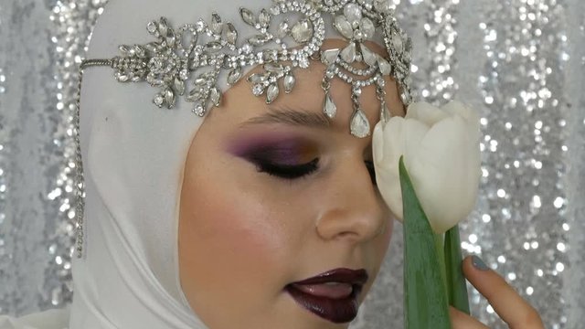 Face of model and tulip in a white image and a silver tiara with a bright multi-colored make-up called a smoky eye, posing in front of the camera in model agency on a silver background. High fashion