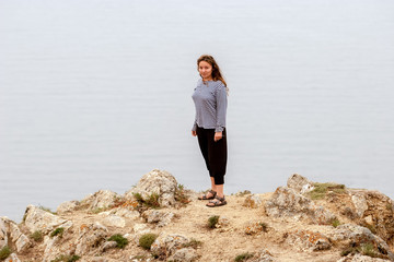 A surprised beautiful girl stands on a rock above the water. European with long dark hair. Singlet, bodysuit and sandals.