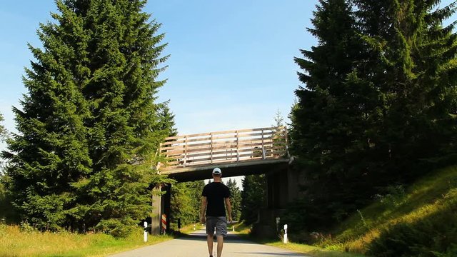 Young man walking on road in white cap and shorts under wood crossover, Sumava National Park, Czech republic and Germany