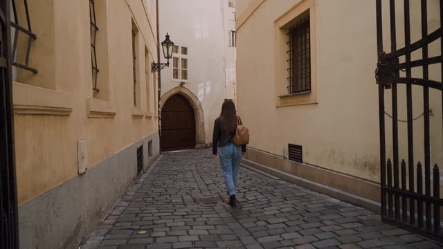 girl in jeans and with backpack is walking along beautiful narrow old street. There is street lamp on wall, arch which is visible in distance. Back view