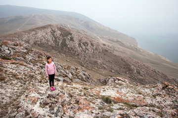 Fototapeta premium A young girl tourist stands on the edge of a high cliff on the shore of Lake Baikal. Girl looking at the camera. The cliffs are red covered with moss. The weather is foggy.