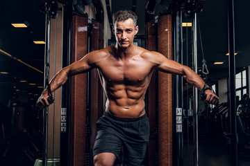 Fototapeta na wymiar Muscular shirtless man is doing exercises with training apparatus at dark gym club.