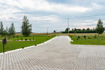Entry for cars into the property made of openwork concrete pavement filled with colorful pebbles, around is green grass and small trees. - obrazy, fototapety, plakaty