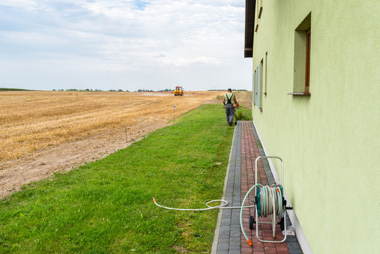 A Man Mowing The Lawn With A Grass Trimmer, Along The Facade Of The House, He Can Be Seen From Behind. A Coiled Garden Hose Is Visible.