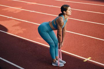 Obraz premium Side view of attractive sporty girl in stylish sportswear stretching with rubber band during workout on stadium
