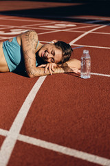 Joyful athlete girl in sportswear happily lying on runner track with bottle after workout on city stadium