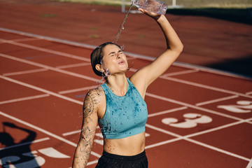 Tired sporty girl in top pouring fresh water on herself after workout on stadium