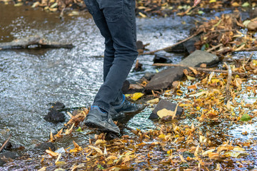 Junge mit schwarzer Hose und Wanderschuhen überquert einen Fluss über einen kleinen Damm aus Steinen und balanciert über glitschige Steine und Blätter und bekommt nasse Füße aber hat draußen viel Spaß