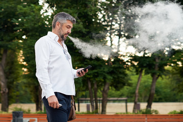 Middle-aged bearded thinking serious concentrated business man standing and smoking electronic cigarette. Blows smoke through the mouth. In-ear wireless earphone. Office centre background