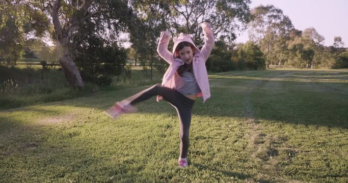 young girl wearing a pink hoodie practices ninja moves, ninjutsu, and tries to hold a pose before running towards the camera arms outstretched in a park in the afternoon golden glow of the setting sun