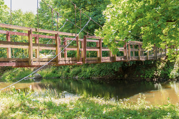 A small wooden suspension bridge passing through the river in the forest, which is mounted on cables against the background of deciduous trees.
