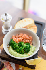 Raw salmon tartare with blanched broccoli and avocado guacamole in bowl on wooden board