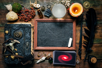 Blackboard with a copy space, tarot cards, crystal ball, magic book and magical wand on the wooden table background. School of magic concept.
