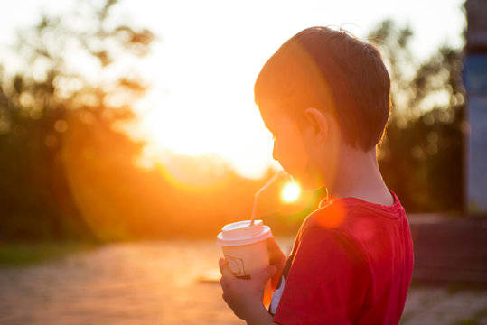 Kid Boy Drinks Cocoa Or Soda In A Cup With Tube On Sunset Background. Silhouette Of A Drinking Boy At Sunset