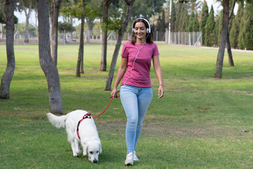 smiling young woman listening to music with her smarthphone and white headphones walking her dog through the park