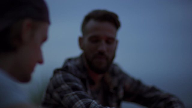 Rack focus of two young male friends talking sitting together outdoors at night