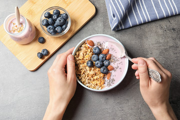 Woman eating tasty yogurt with oatmeal and blueberries at grey table, top view