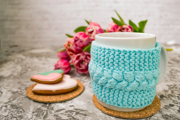 White cup of tea in a knitted blue case with pink flowers tulips and sweets, with gingerbread on a textural table.
