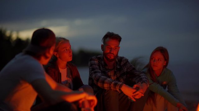 Tilt up shot of four young adventure seekers sitting together by campfire at night and talking