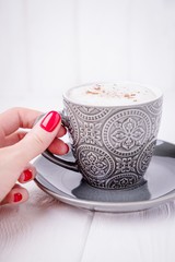 Female hand holding gray cup of coffee cappuccino with cinnamon. Manicure with red nail polish. Copy space