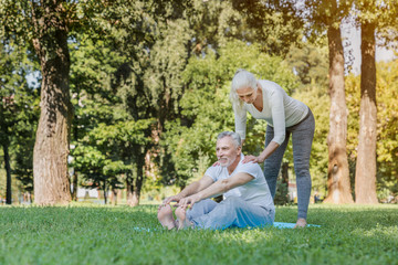 Naklejka na ściany i meble Senior woman helping to stretch mature man outdoors at park while doing workout and yoga exercices