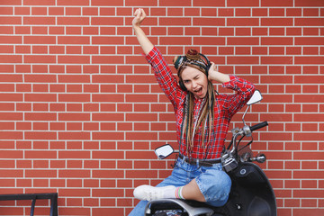Fototapeta na wymiar Happy young hipster woman listen to music and rise hand up sitting on moped