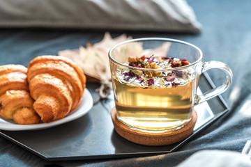 Cozy autumn morning concept. Breakfast with herbal tea and croissants on the bed with grey sheets in a sunny room