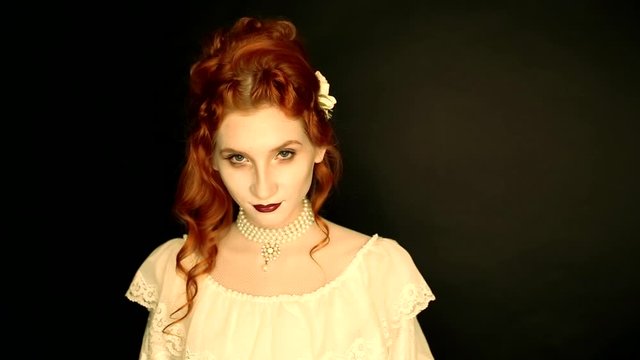Young vampire woman with long curly hair, pale skin in a white dress on a black background. A beautiful redhead model with red lips. Outfit for halloween. Vampire with open mouth and fangs