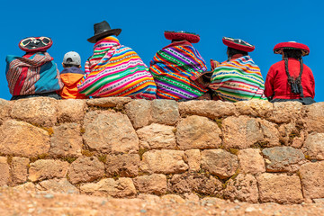 A group of Quechua indigenous women in traditional clothing and a young boy sitting and chatting on an ancient Inca wall in the archaeological site of Chinchero in the region of Cusco city, Peru. - obrazy, fototapety, plakaty