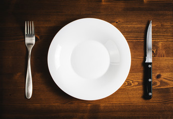 White empty plate with a fork and knife on a wooden table.