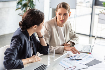 two businesswomen in formal wear at table looking at each other in office
